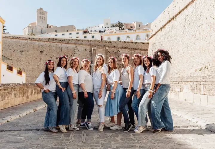 Dix amies célèbrent leur evjf en France, souriantes et parées de couronnes de fleurs, devant un mur de pierre et des bâtiments blancs.