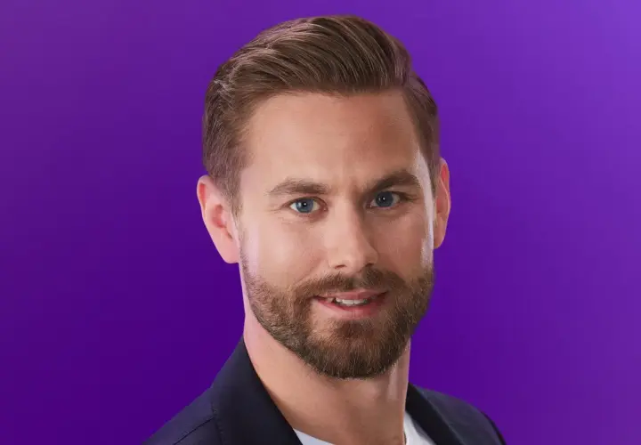 Oskar från Love is Blind, med blå ögon och skägg, ler mot kameran mot en lila bakgrund.