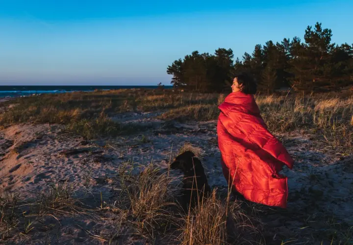 Kobieta w czerwonym śpiworze i pies na plaży. Zastanawiasz się, jaki śpiwór wybrać na nocne wędrówki?