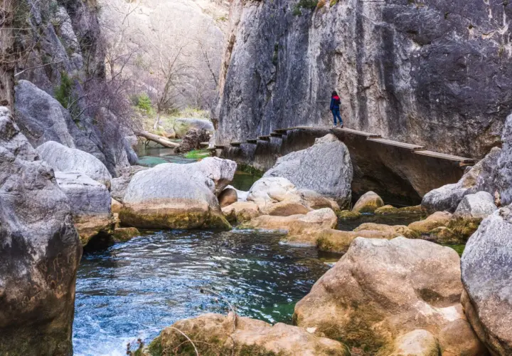 Una persona camina por un puente de madera en la ruta del silencio, rodeada de rocas y un río de aguas cristalinas.
