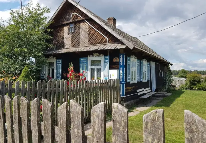 Drewniany dom z niebieskimi, otwartymi okiennicami, ozdobiony ludowymi wzorami. Wokół zielony trawnik i drewniany płot.