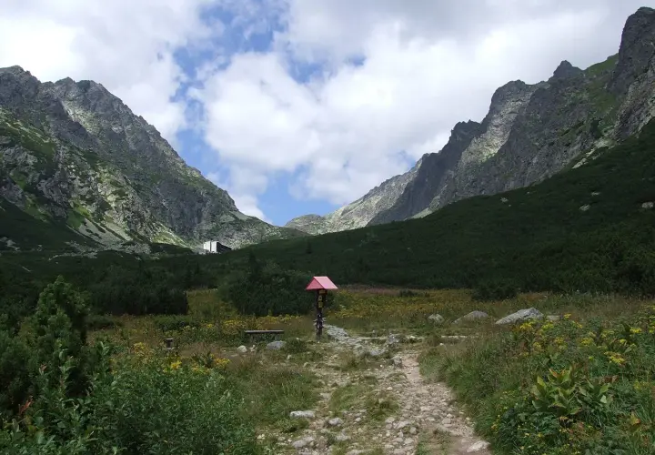 Kamienista ścieżka w górskiej dolinie z zielonymi zboczami i budynkiem w tle.