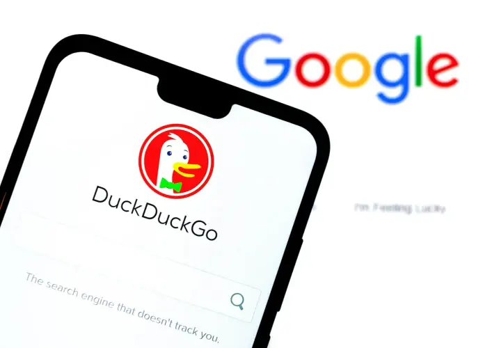 Na ekranie telefonu widoczny jest interfejs wyszukiwarki DuckDuckGo, która jest najlepszą wyszukiwarką internetową dbającą o prywatność. W tle rozmyty napis Google.
