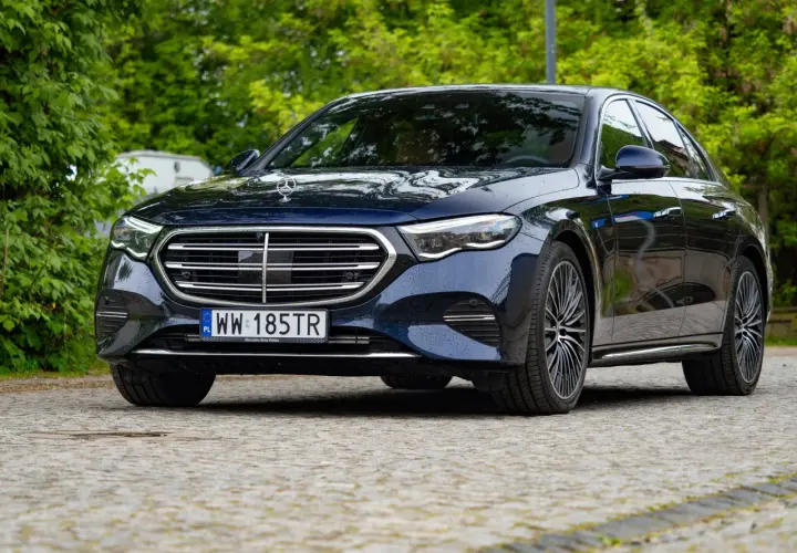 Elegancki, ciemnogranatowy Mercedes z napędem 4matic, zaparkowany na brukowanej nawierzchni, otoczony zielenią.