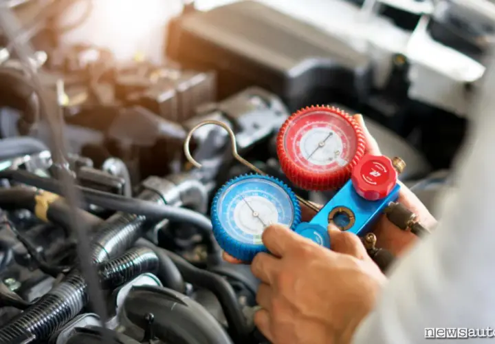 Manometro per ricaricare aria condizionata auto, con due quadranti (rosso e blu) collegati a tubi, in mano a un meccanico davanti al motore.