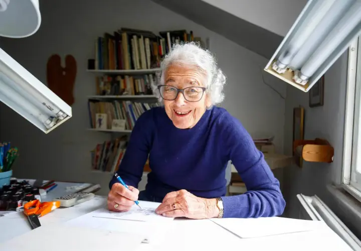 Judith Kerr, lächelnd, zeichnet an ihrem Schreibtisch. Bücherregale im Hintergrund.