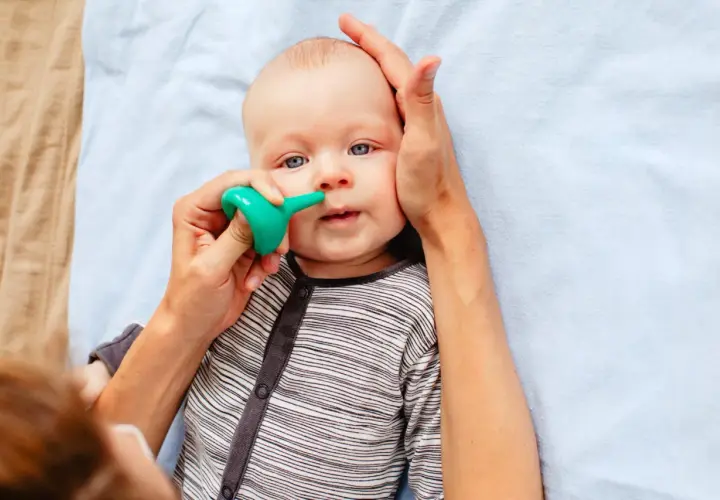 Un bebé con ojos azules recibe un lavado nasal con un aspirador verde. Una mano sujeta su cabeza mientras la otra usa el dispositivo.