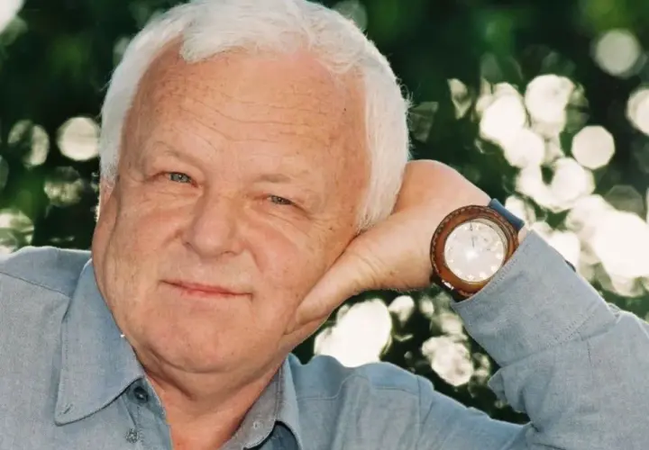 Uśmiechnięty Marian Opaniak, z siwymi włosami i niebieskimi oczami, opiera głowę na dłoni. Zegarek na jego nadgarstku wskazuje godzinę.