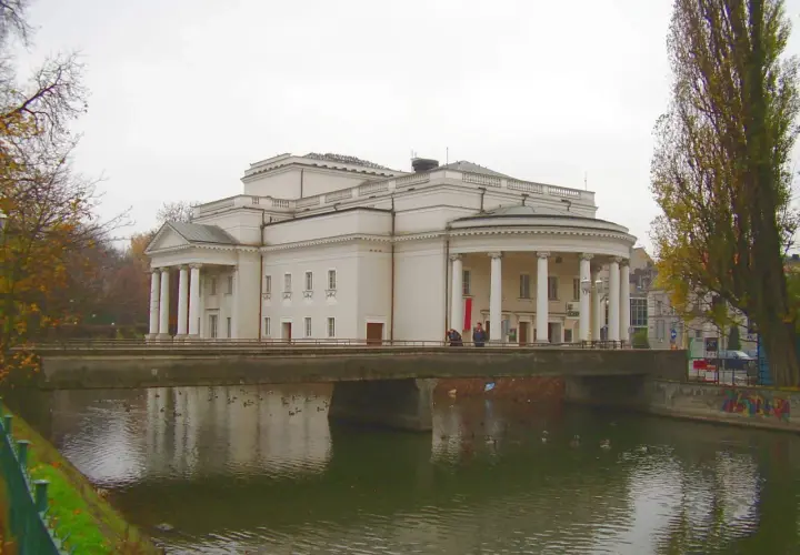 Teatr Kalisz zaprasza na spektakle. Imponujący budynek z kolumnadą nad rzeką, jesienne drzewa.