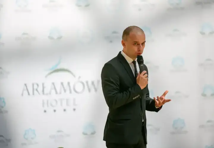 Mężczyzna w garniturze przemawia na tle logo Hotel Arłamów. Właściciel hotelu z pasją opowiada o swoich planach.