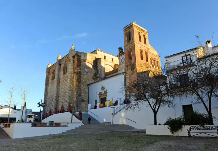 Iglesia de la Asunción en Cazalla de la Sierra, con su torre campanario y fachadas de piedra y cal bajo un cielo azul.
