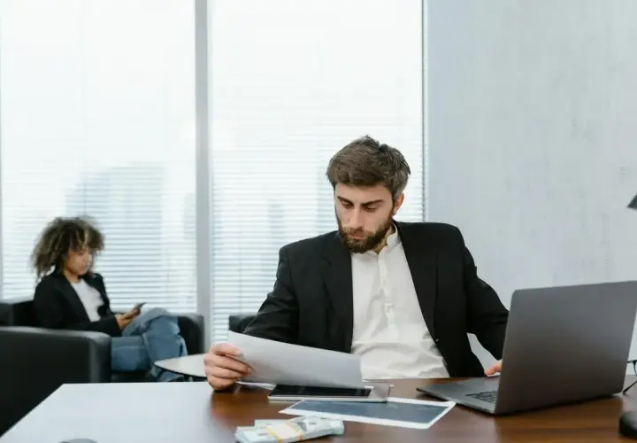 A man in a suit reviews documents, perhaps researching accelerated online MPA programs. A woman in the background is on her phone.