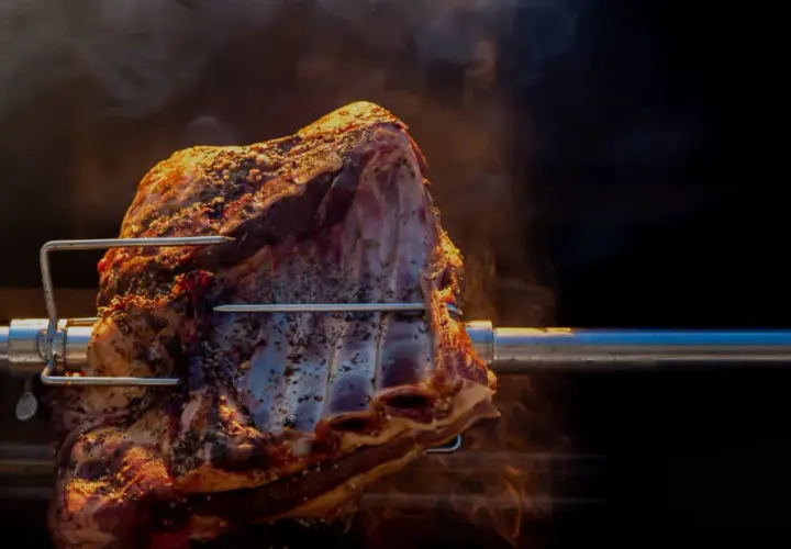 Un morceau de viande assaisonné tourne lentement sur une broche, dégageant une fumée alléchante. La cuisson à la broche promet un délice savoureux.