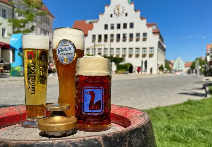 Drei Gläser Bier, darunter ein helles und ein dunkles, stehen auf einem Fass. Im Hintergrund eine malerische Stadtkulisse mit einem historischen Gebäude. Ein perfekter Genuss von italienischem Bier.