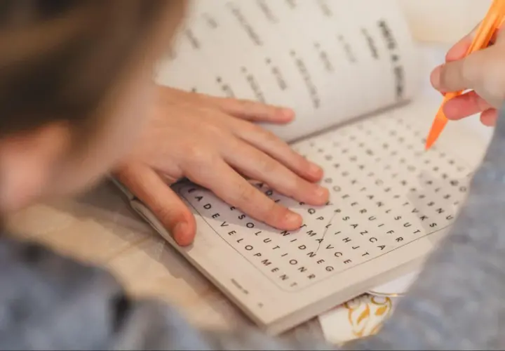 Mano de niño resolviendo una sopa de letras para la memoria con un lápiz naranja.
