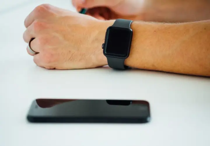 Czarny smartwatch na nadgarstku i leżący obok smartfon. Dowiedz się, jak włączyć bluetooth w zegarku, by połączyć go z telefonem.