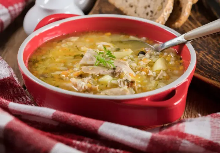 Gorąca zupa krupnik przepis babci w czerwonej misce, z kawałkami mięsa, ziemniaków i kaszy pęczak. Obok kromki chleba i czerwono-biała serwetka.