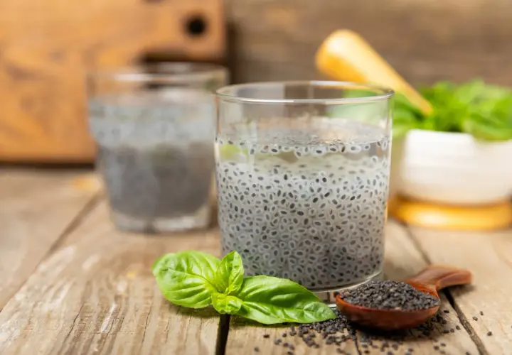 Due bicchieri con semi di basilico in acqua, un cucchiaino di semi e foglie fresche su un tavolo di legno.
