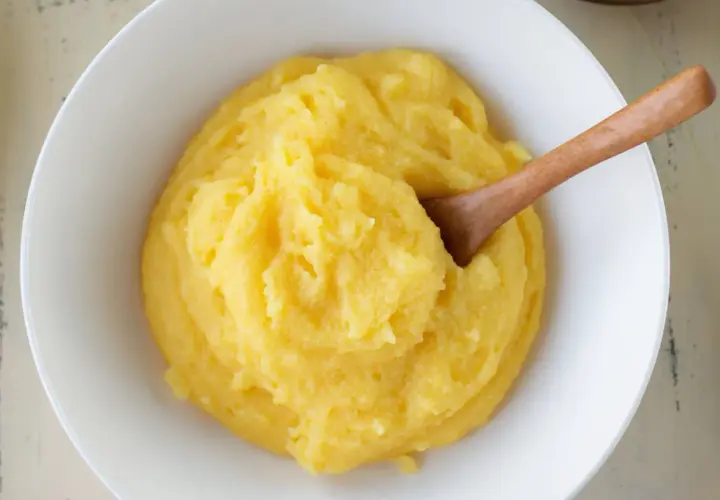 Un tazón blanco lleno de polenta cremosa, con una cuchara de madera. ¿Sabes qué es la polenta? Es un plato reconfortante.