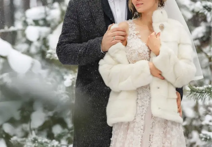 Para młoda w zimowej scenerii. Ona w białym futerku i koronkowej sukni, on w eleganckim płaszczu. Ich ślub zimą wygląda bajkowo.