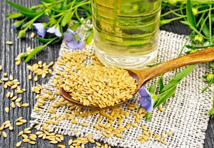 Semi di lino dorati su un cucchiaio di legno, con una bottiglia di olio di lino e fiori di lino sullo sfondo.