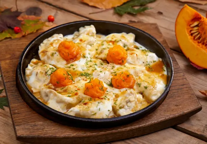 Pierożki z dynią w sosie, posypane koperkiem, podane w żeliwnej patelni na drewnianym blacie. Obok kawałek dyni.