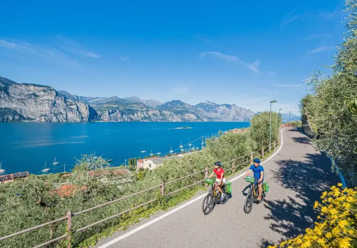 Two people enjoy biking along a scenic road in Italy, with a stunning blue lake and mountains in the background.