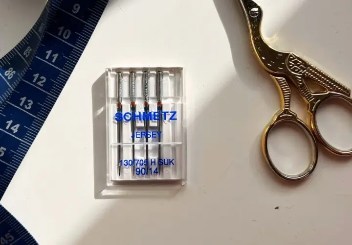 Schmetz Jersey needles, size 90/14, are ideal for stretch fabrics. A blue measuring tape and gold scissors complete the sewing setup.