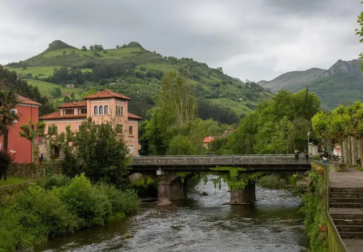 Un puente cruza un río, conectando pueblos bonitos cerca de Liérganes. Edificios rosados y verdes colinas completan la escena.
