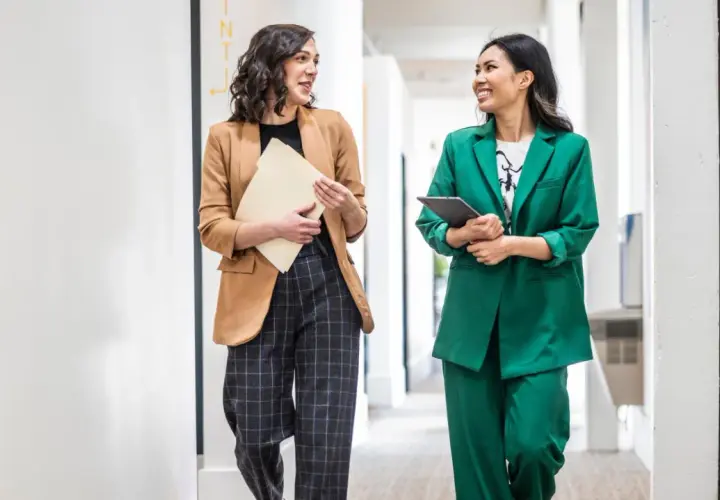 Zwei Frauen in lässiger Business-Kleidung gehen durch einen Flur. Eine trägt einen braunen Blazer und karierte Hose, die andere einen grünen Hosenanzug.