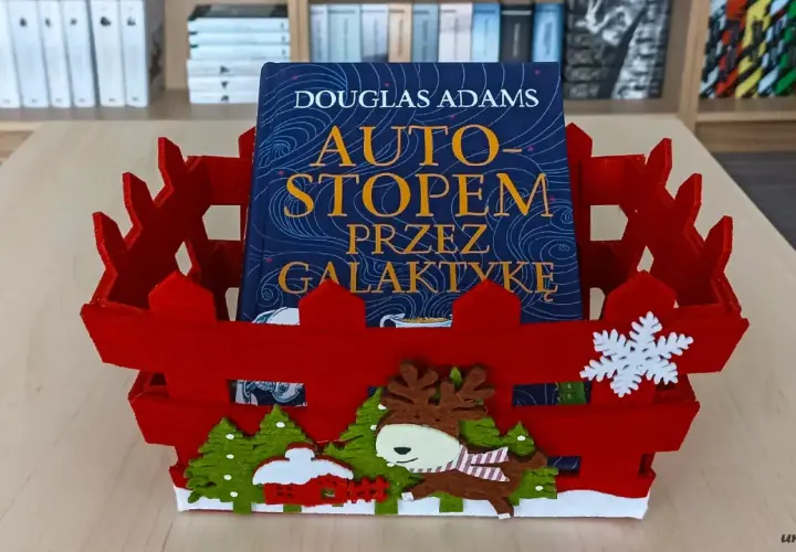 Książka "Autostopem przez galaktykę" Douglasa Adamsa w czerwznym, świątecznym koszyku z reniferem i choinkami.