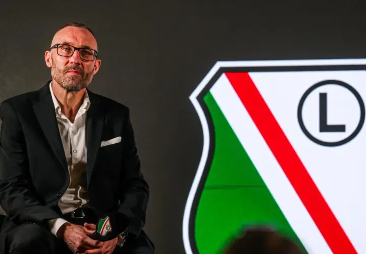 Mężczyzna w okularach, w garniturze, siedzi obok herbu Legii Warszawa. To nowy trener Legii Warszawa.