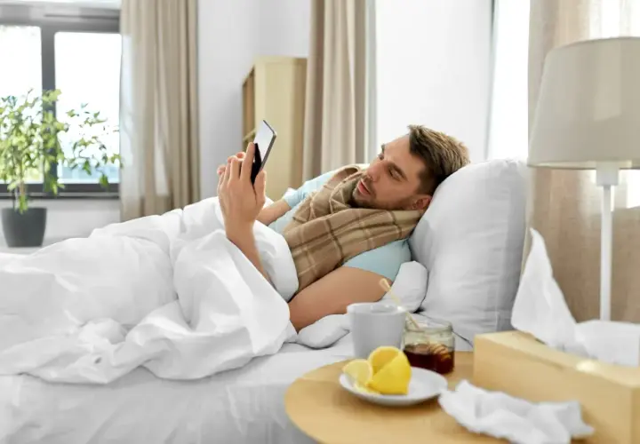 Chory mężczyzna w łóżku, owinięty szalikiem, przegląda telefon. Obok kubek z herbatą, cytryna i miód. Czyżby czekał na zwolnienie lekarskie z powodu siły wyższej, zgodnie z kodeksem pracy?