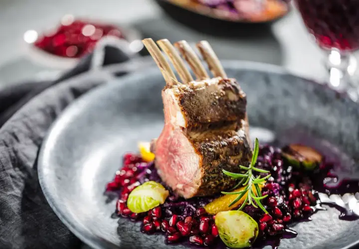 Carré d'agneau parfaitement rôti, servi avec une garniture colorée de grenade, chou rouge et romarin. La cuisson du carré d'agneau est mise en valeur par la présentation.