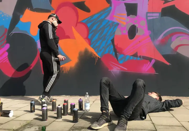 Zwei Künstler, eine Grafikdesignerin und ihr Kollege, sprühen Graffiti. Einer liegt entspannt, der andere sprüht Farbe auf die Wand.