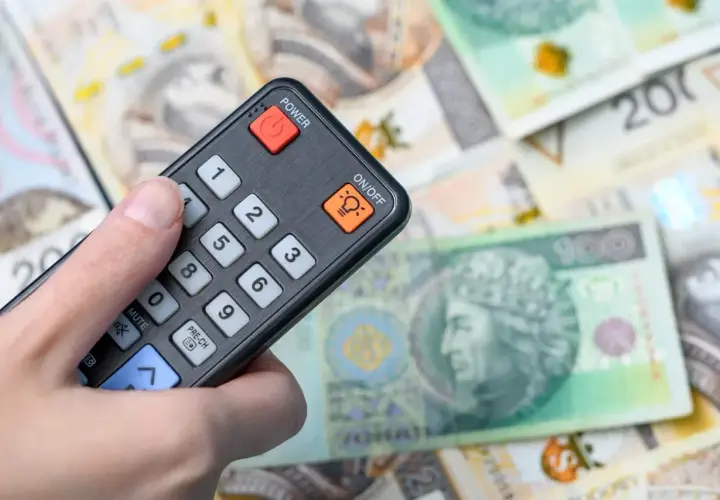Dłoń trzyma pilot na tle polskich banknotów. Zastanawiasz się, ile kosztuje abonament RTV?