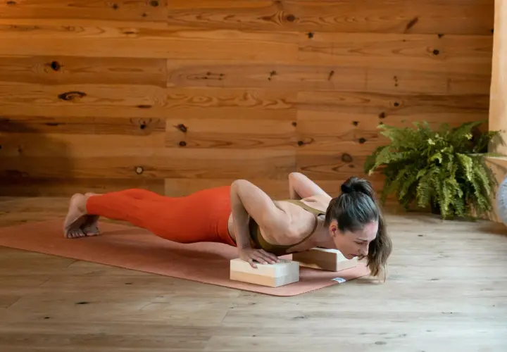 Kobieta w pomarańczowych legginsach wykonuje pozycję chaturanga dandasana, opierając dłonie na drewnianych klockach.