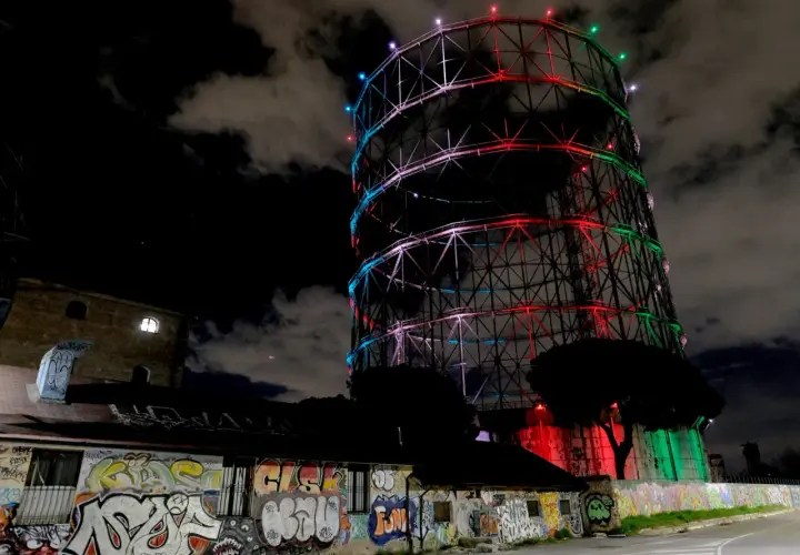 Il gasometro di Roma, illuminato da luci colorate, si staglia contro il cielo notturno. Un muro di graffiti decora l'edificio sottostante.