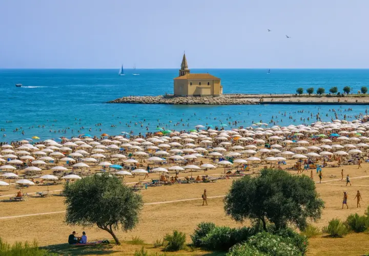 Vue aérienne d'une plage bondée à Caorle, avec des rangées de parasols blancs, des baigneurs dans la mer bleue et une petite église sur un îlot rocheux.