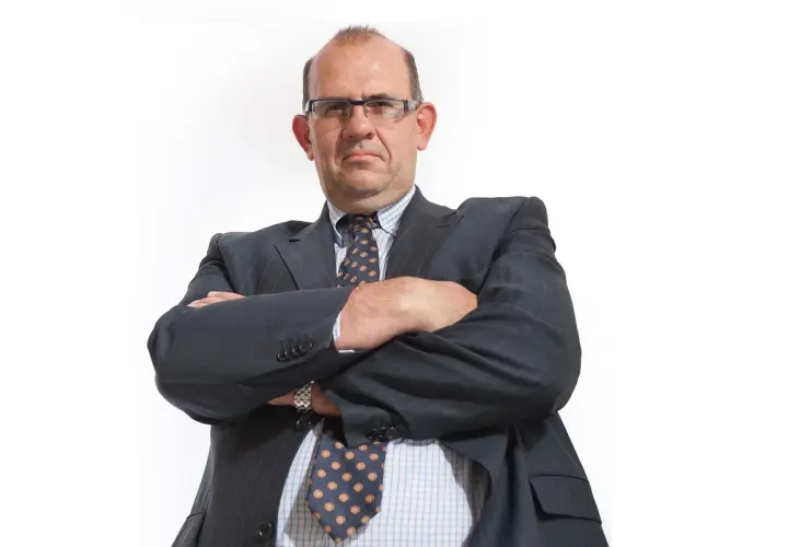 Mężczyzna w garniturze i okularach ze skrzyżowanymi ramionami, patrzący na wprost.