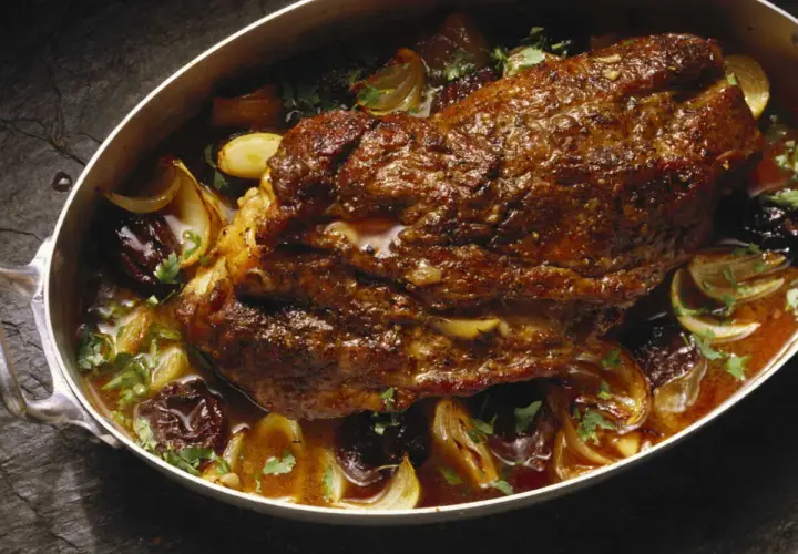 Rouelle de porc rôtie dans un plat avec des oignons caramélisés, des pruneaux et du persil frais.