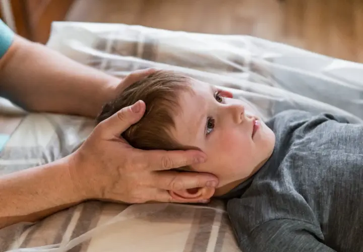 Chłopiec z obniżonym napięciem mięśniowym leży na plecach, a terapeuta delikatnie dotyka jego głowy.