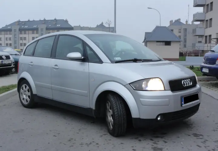 Srebrne Audi A2 z włączonymi światłami, zaparkowane na tle nowoczesnych budynków mieszkalnych.