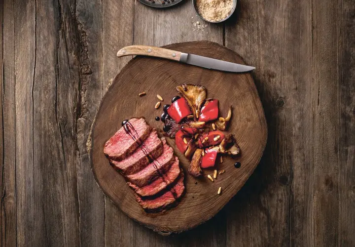 Saftige Tagliata di Manzo mit gebratenen Pilzen und Paprika auf einem rustikalen Holzbrett, garniert mit Pinienkernen und Balsamico.