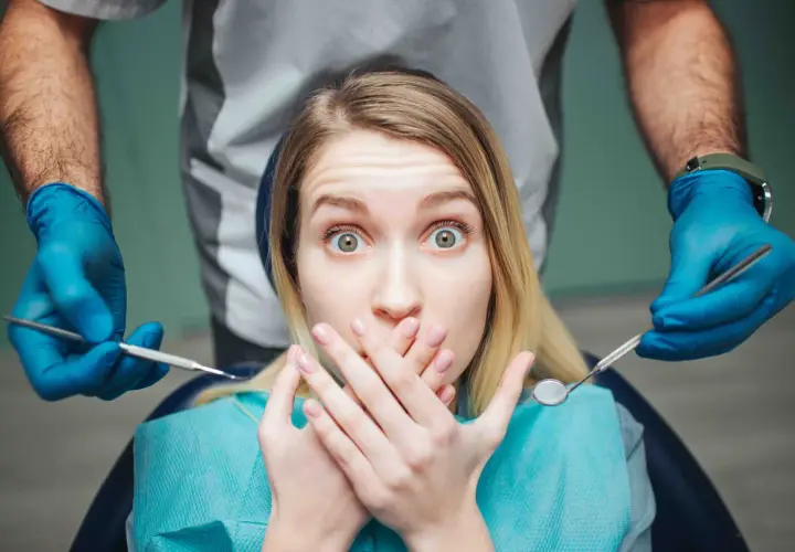 Occhi sbarrati e mani sulla bocca: la paura del dentista è palpabile mentre il dentista si avvicina con gli strumenti.