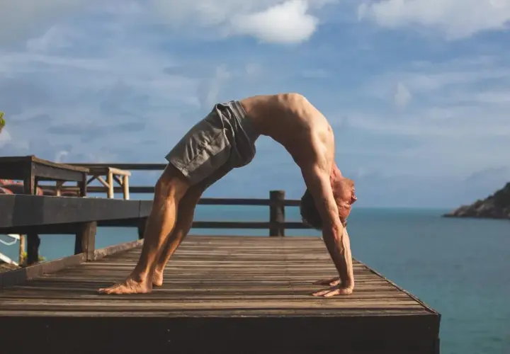 Mężczyzna wykonuje pozycję urdhva dhanurasana na drewnianym tarasie z widokiem na ocean i niebo.