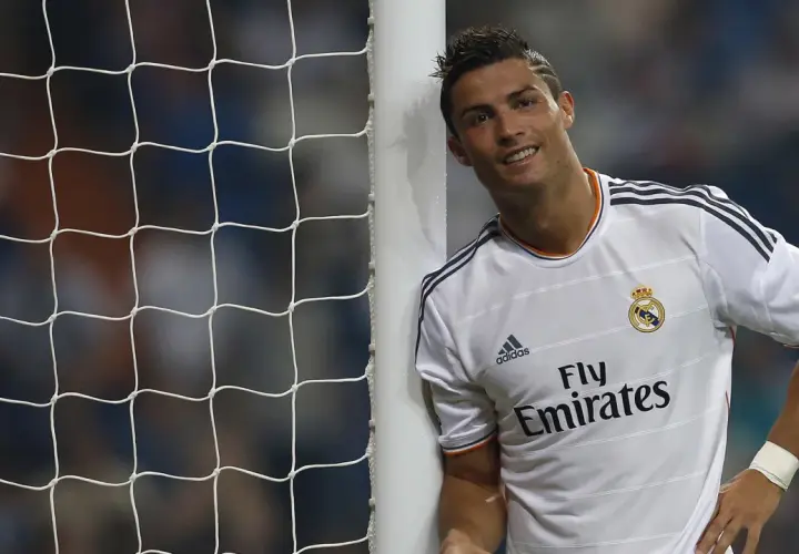 Uśmiechnięty Cristiano Ronaldo opiera się o słupek bramki. Jego sylwetka i sportowa forma, choć nie widać tu konkretnych danych o Ronaldo Wzrost i Waga, świadczą o jego atletyzmie.