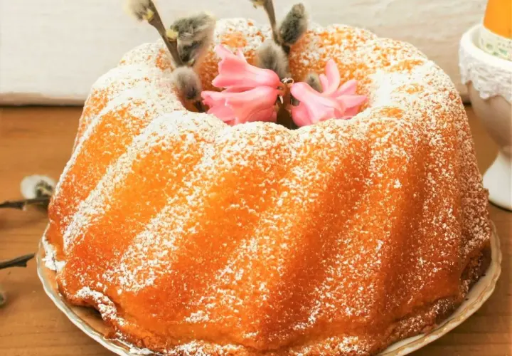 Puszysta babka jogurtowa oprószona cukrem pudrem, ozdobiona baziami i różowymi kwiatami. Idealna na wielkanocny stół.