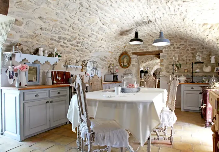 Cuisine rustique au style shabby chic, avec murs en pierre, table dressée et chaises anciennes.
