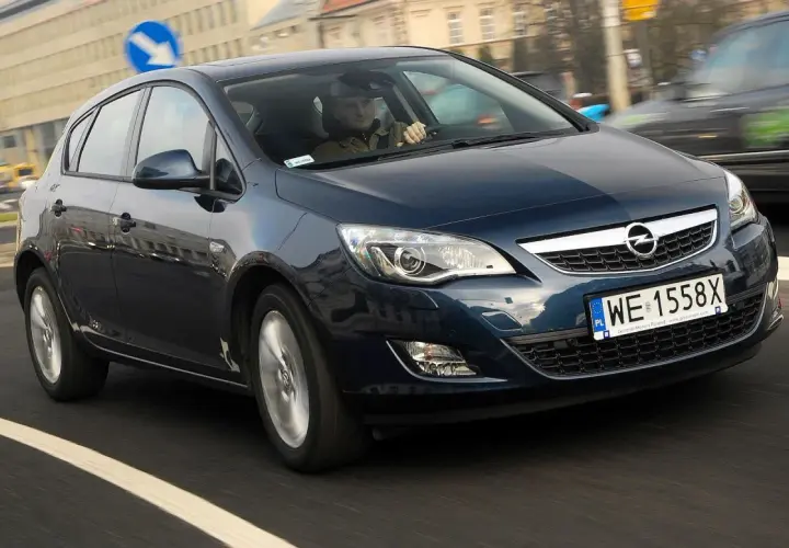 Ciemnoniebieski Opel Astra J 1.6 benzyna w ruchu miejskim. Kierowca w czapce prowadzi auto.